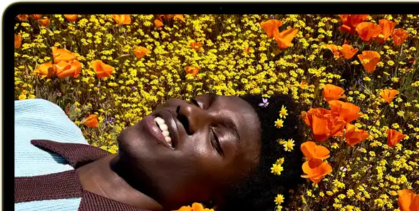 MacBook Neo, Farbe Zitrus, das Display zeigt eine Person, die in einer Blumenwiese in der Sonne liegt und lächelt